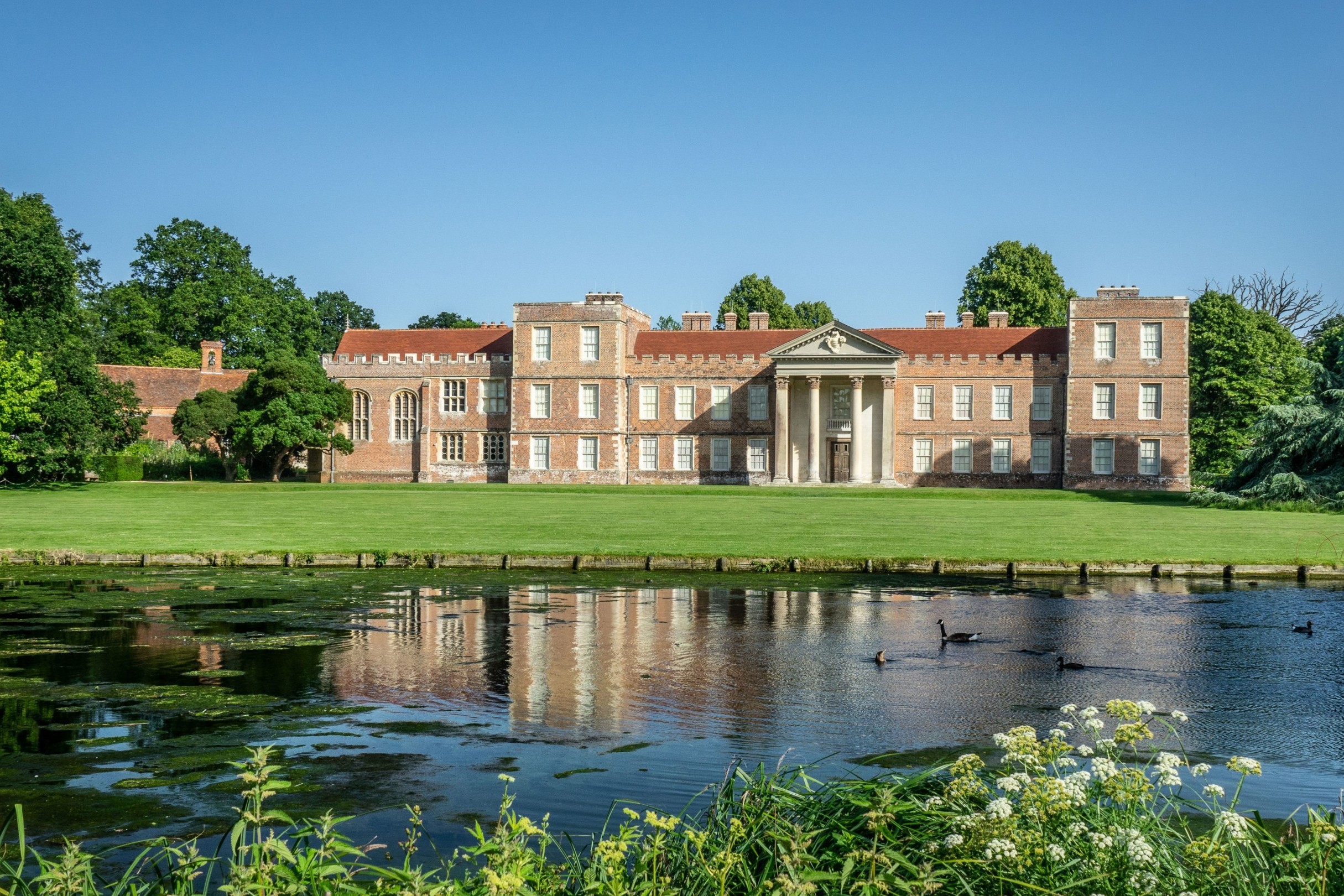 The Vyne country house reflected in a lake, surrounded by lawns and trees on a sunny day.