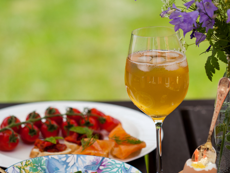 A glass of wine with ice in it, alongside some food on a wooden table, outside in a garden.