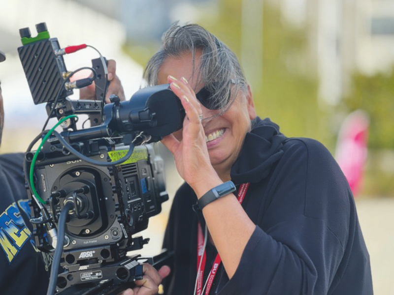 A smiling woman wearing a black shirt is looking through a viewfinder of a large film camera