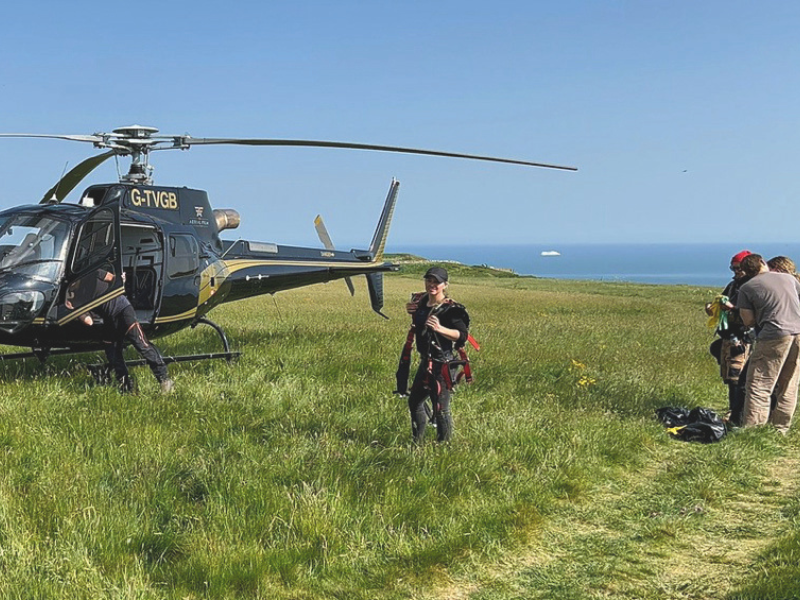 A helicopter on an elevated field overlooking the Isle of Wight coastline, there are people standing nearby as part of a film shoot