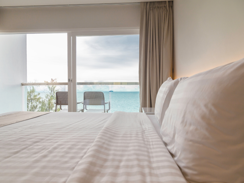 A hotel bed with crisp white linen, gives way to a balcony overlooking the sea, through floor-to-ceiling glass doors