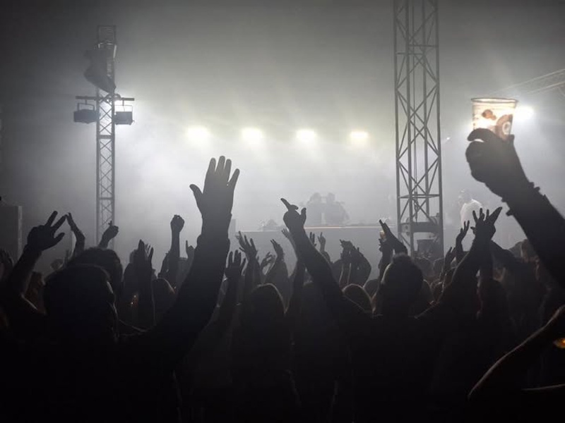 A crowd of people in a music tent in silhouette, hands in the air against the backdrop of spotlights and acts on stage