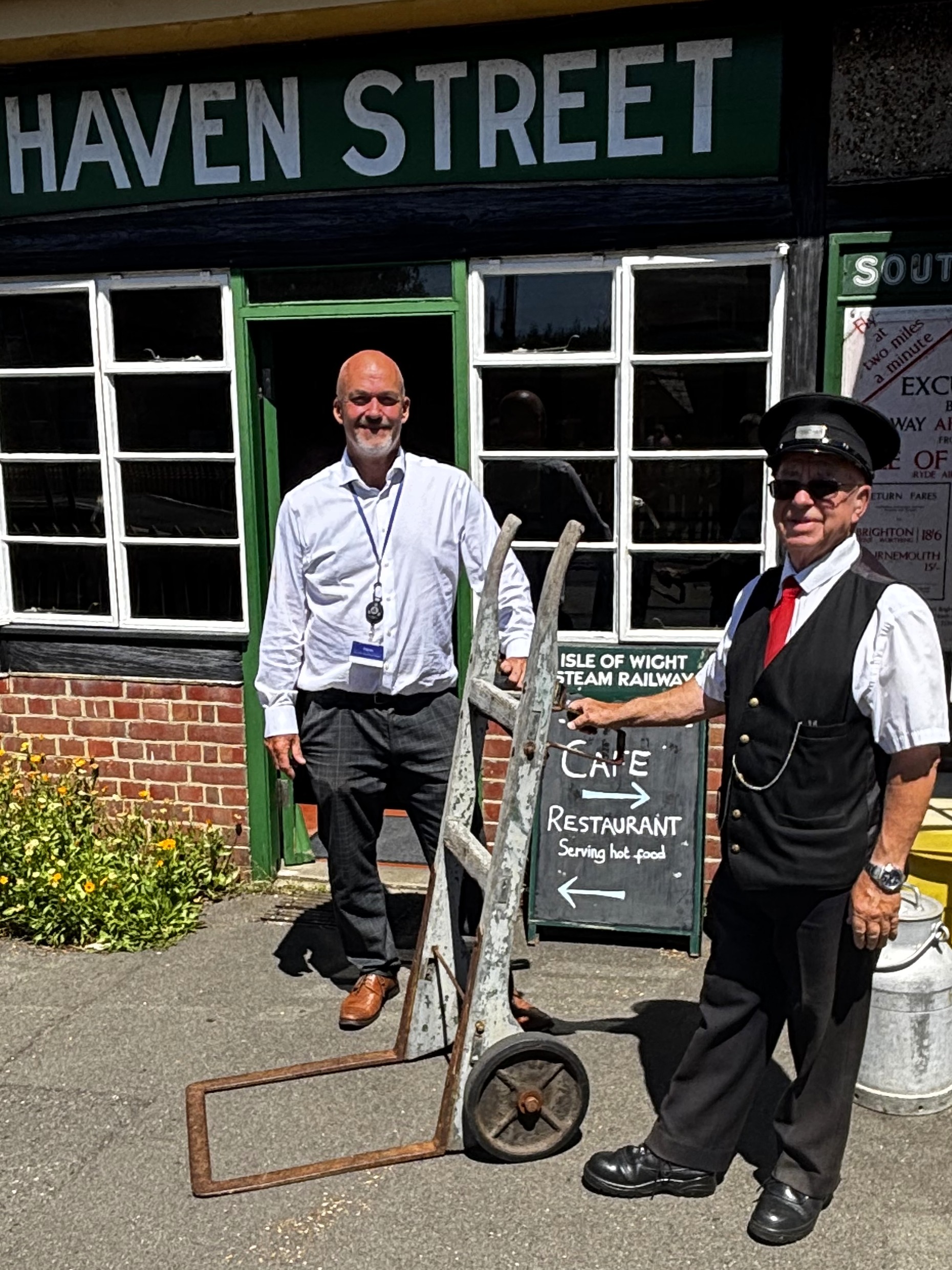Two people (Wightlink’s Martin Gulliver and Isle of Wight Steam Railway volunteer Bob Comer) standing outside Haven Street station on the Isle of Wight Steam Railway beside a historic metal luggage trolley. The station building features green-and-white signage reading “HAVEN STREET”, with a café and restaurant sign visible near the entrance. The scene is photographed on a bright sunny day.