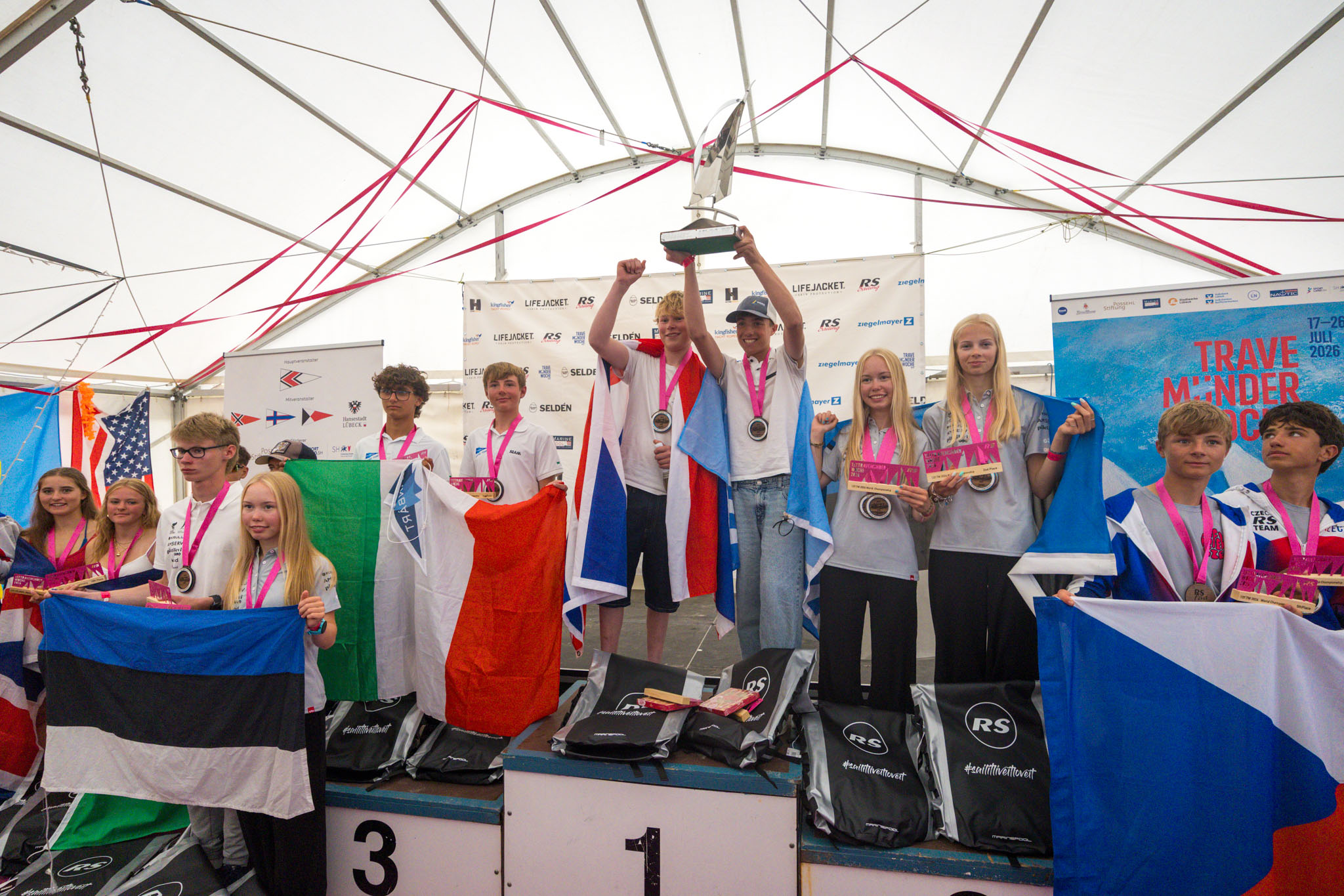 Young sailors standing on a winners’ podium with medals, national flags and a trophy at the RS Feva World Championships.
