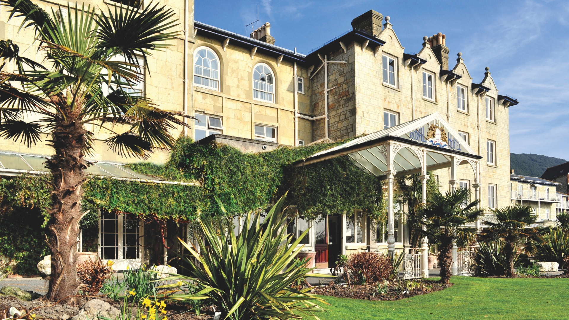 Exterior view of The Royal Hotel, a historic stone building surrounded by landscaped gardens, palm trees and ornamental planting. A covered entrance porch welcomes guests beneath a bright blue sky.
