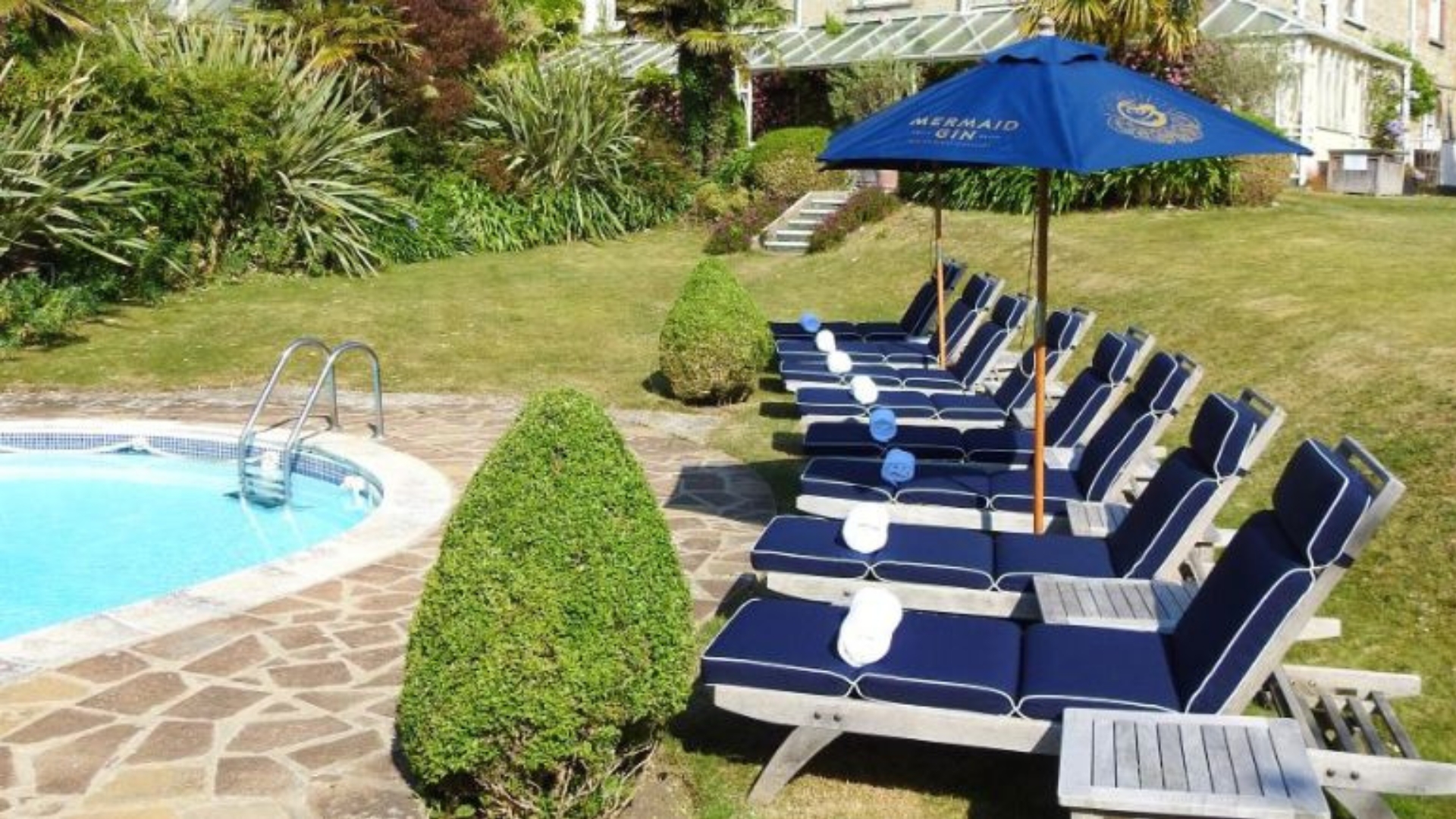Outdoor swimming pool with a row of cushioned sun loungers arranged along the poolside terrace. Manicured gardens, parasols and the hotel building create a relaxing setting for leisure and outdoor dining.