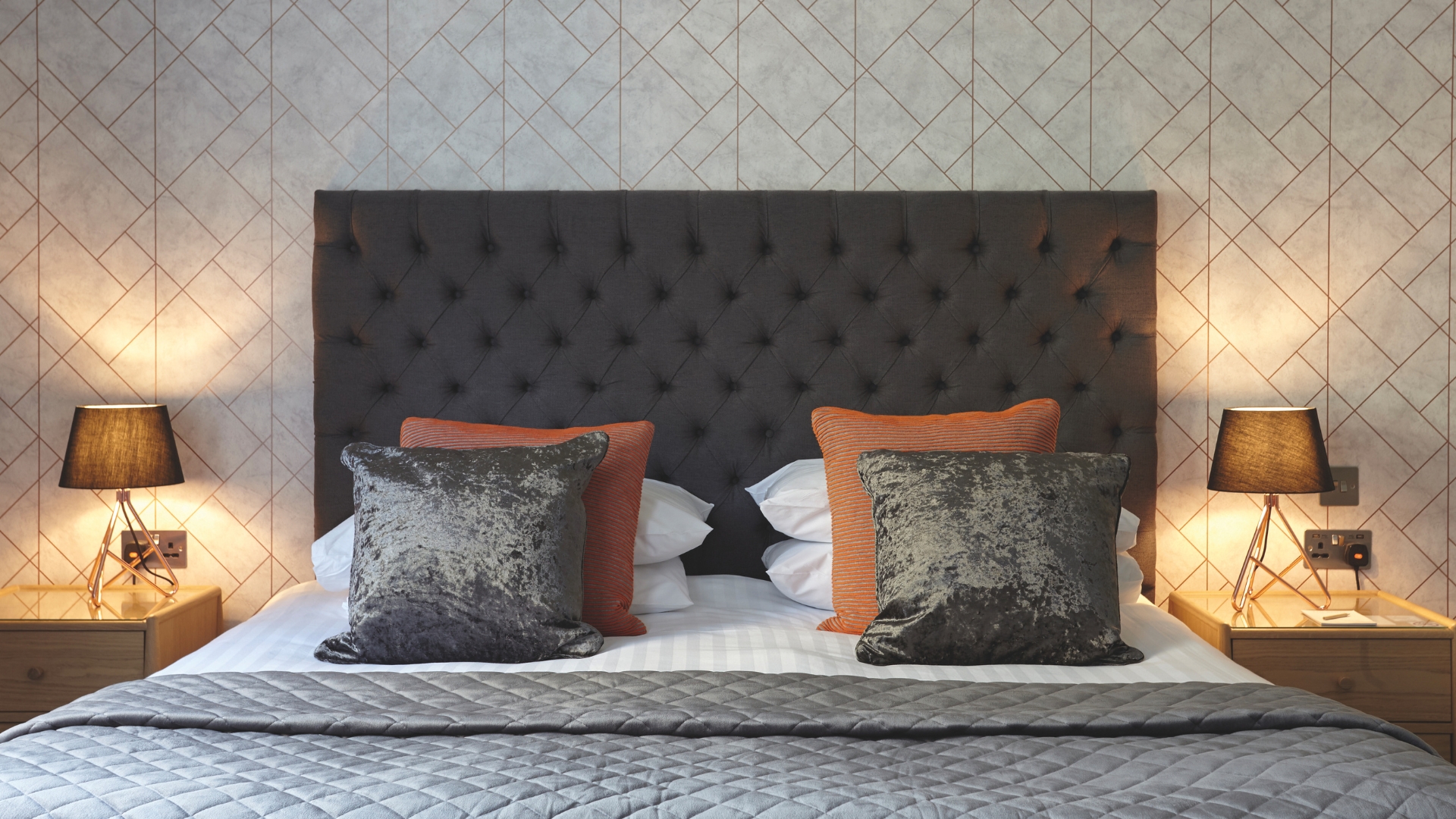 Elegant hotel bedroom featuring a large double bed with a dark upholstered headboard, decorative cushions and matching bedside tables. Warm bedside lamps and geometric-patterned wallpaper create a stylish and comfortable setting.