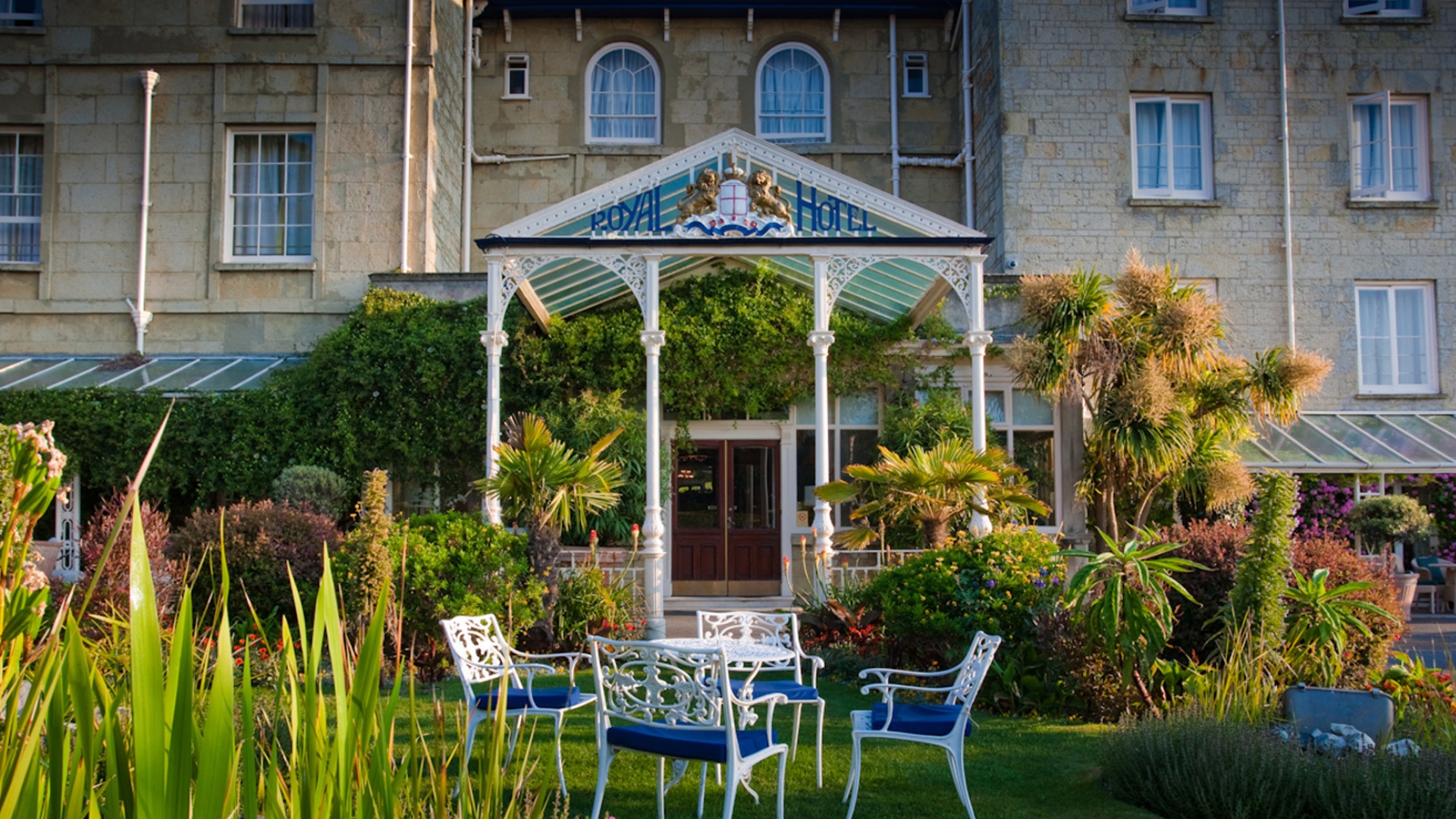 The Royal Hotel on the Isle of Wight, viewed from landscaped gardens featuring ornamental plants, palm trees and white outdoor seating. The historic stone hotel building stands behind an ornate covered entrance with “Royal Hotel” signage, surrounded by colourful planting and manicured lawns in warm sunlight.