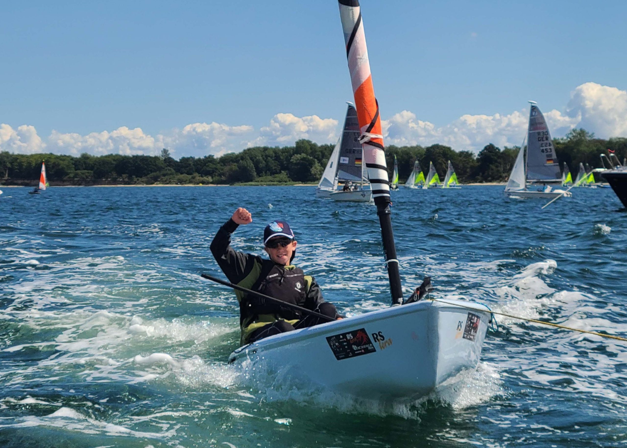 Young sailor in a single-handed dinghy on open water, raising a fist while sailing, with several other sailboats and a tree-lined shoreline in the background.