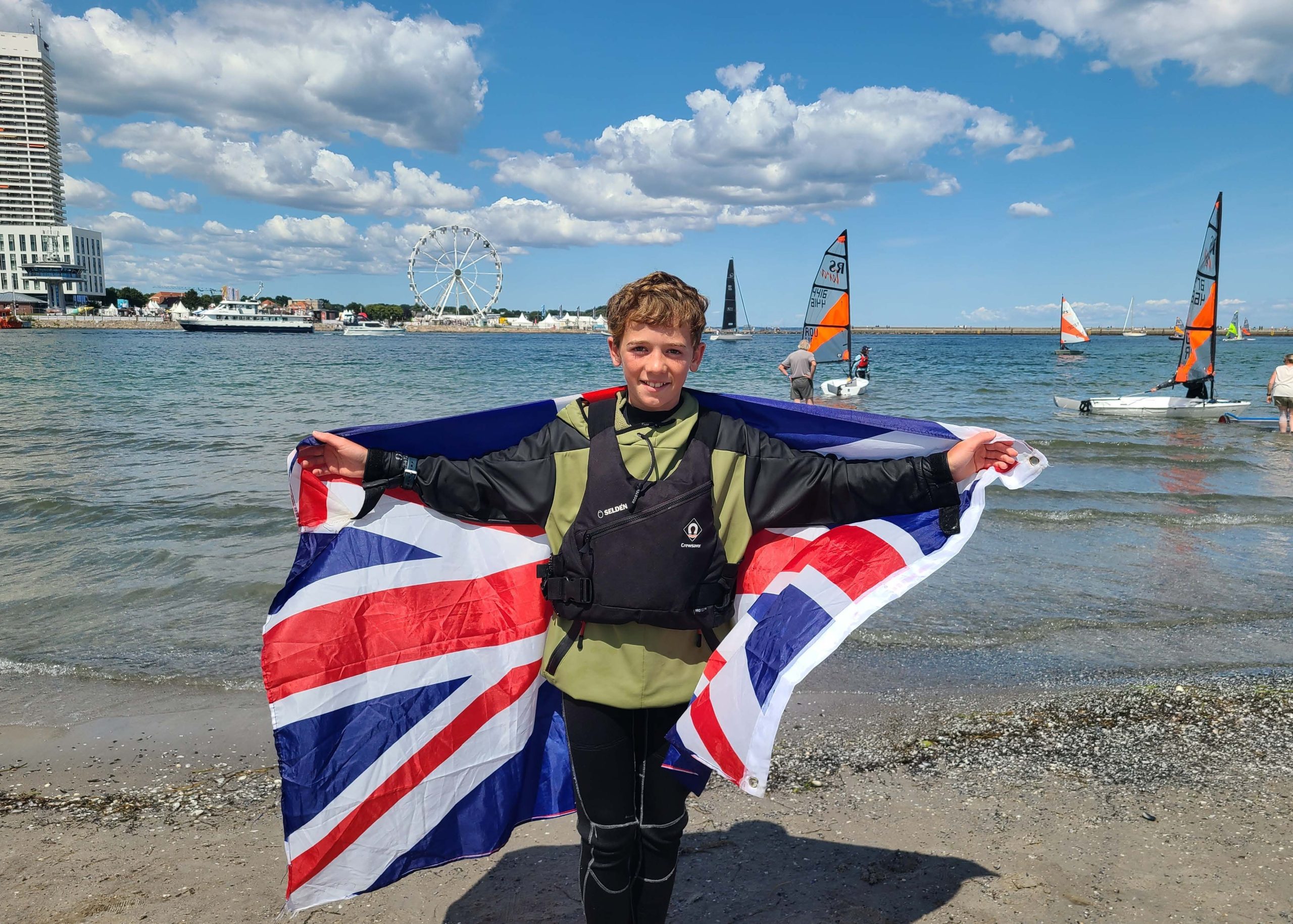 Young sailor standing on a beach holding a large Union Flag, with sailing dinghies on the water and a marina waterfront in the background.