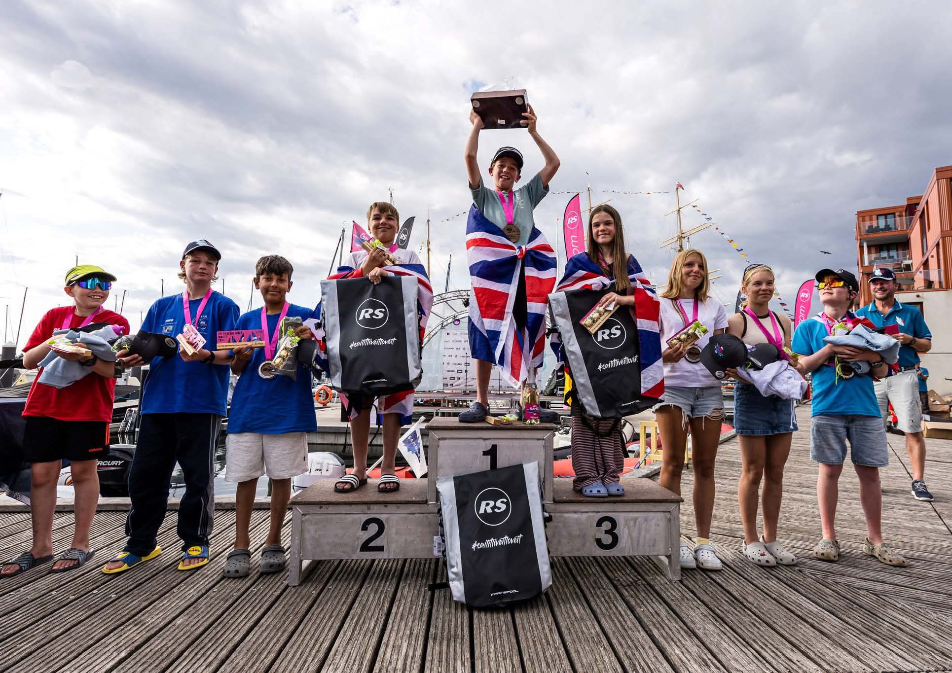 Junior sailors standing on a winners' podium at a marina after a sailing competition, displaying medals, trophies and prize bags, with boats and waterfront buildings behind them.