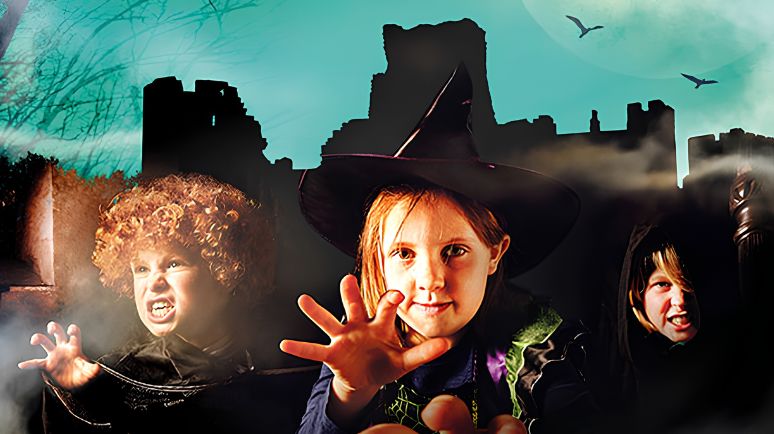 Children in Halloween costumes in front of Carisbrooke Castle ruins with mist, bats and a moonlit sky.