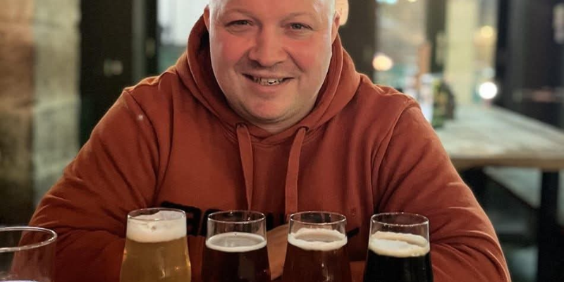 A man wearing a rust coloured hoodie sits in a bar with 4 tasting ales in front of him
