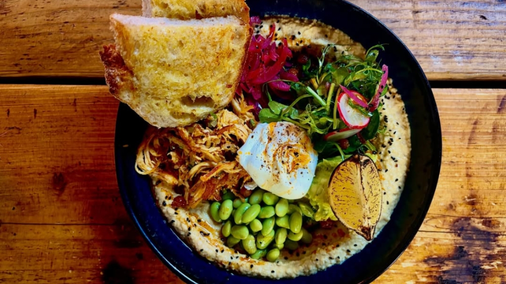 Colourful brunch bowl topped with pulled chicken, a poached egg, edamame beans, pickled vegetables and toasted bread, served in a black bowl on a wooden table.