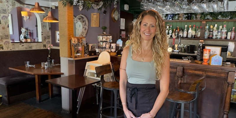 A woman stands in front of a traditional pub bar, smiling at the camera