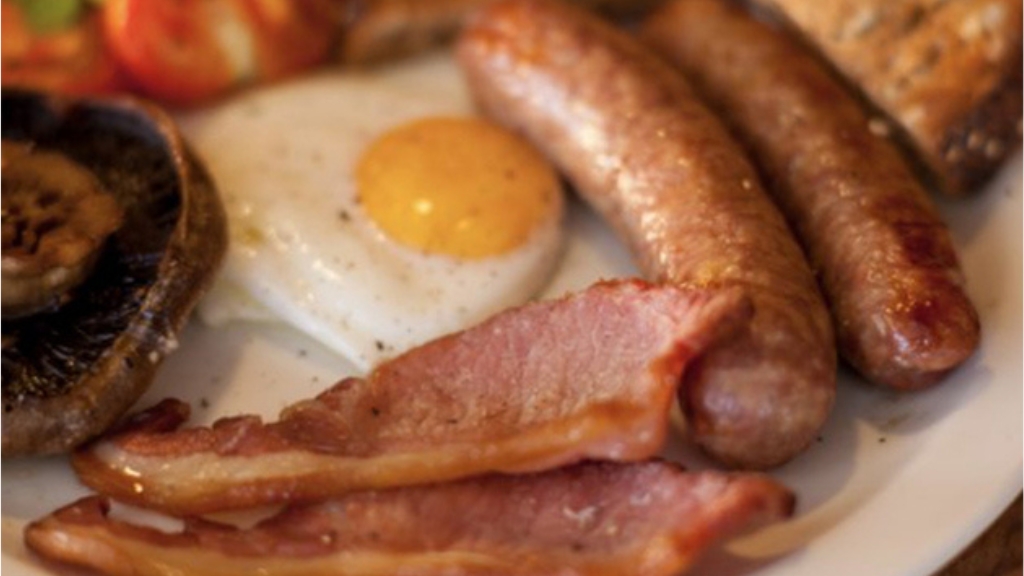 Traditional cooked breakfast featuring sausages, bacon, a fried egg and grilled mushroom served on a white plate.