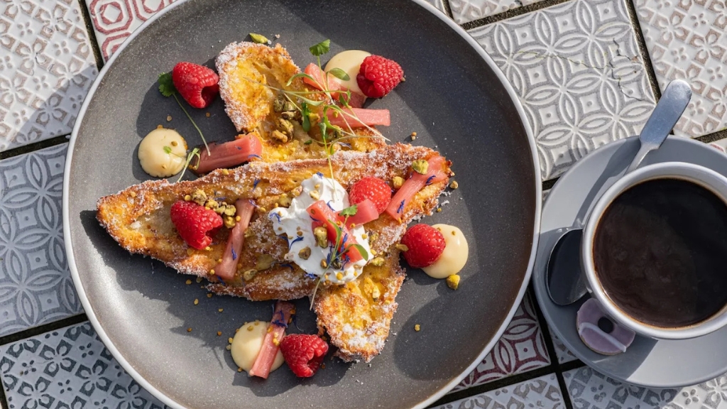 Plate of sweet French toast croissants topped with raspberries, rhubarb, cream and crushed pistachios, served alongside a cup of black coffee on a patterned tiled table.