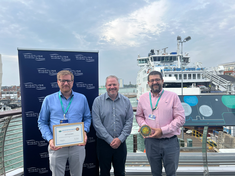 Three people stand on an outdoor ferry terminal balcony in front of a Wightlink-branded backdrop and a docked ferry. The person on the left holds a framed certificate, while the person on the right holds a round award. The harbour, ferry infrastructure and a cloudy sky are visible in the background.