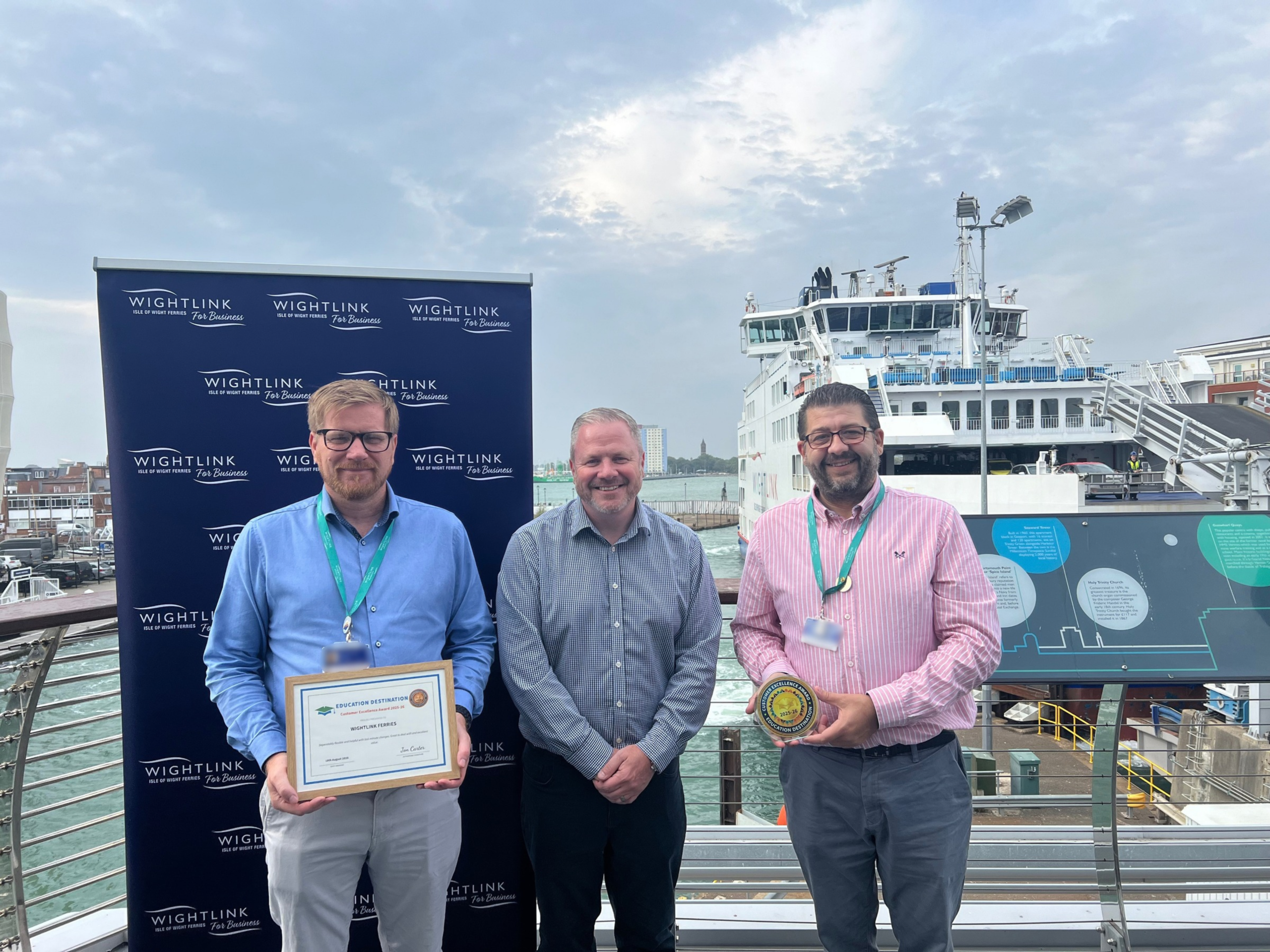 Three people stand on an outdoor ferry terminal balcony in front of a Wightlink-branded backdrop and a docked ferry. The person on the left holds a framed certificate, while the person on the right holds a round award. The harbour, ferry infrastructure and a cloudy sky are visible in the background.