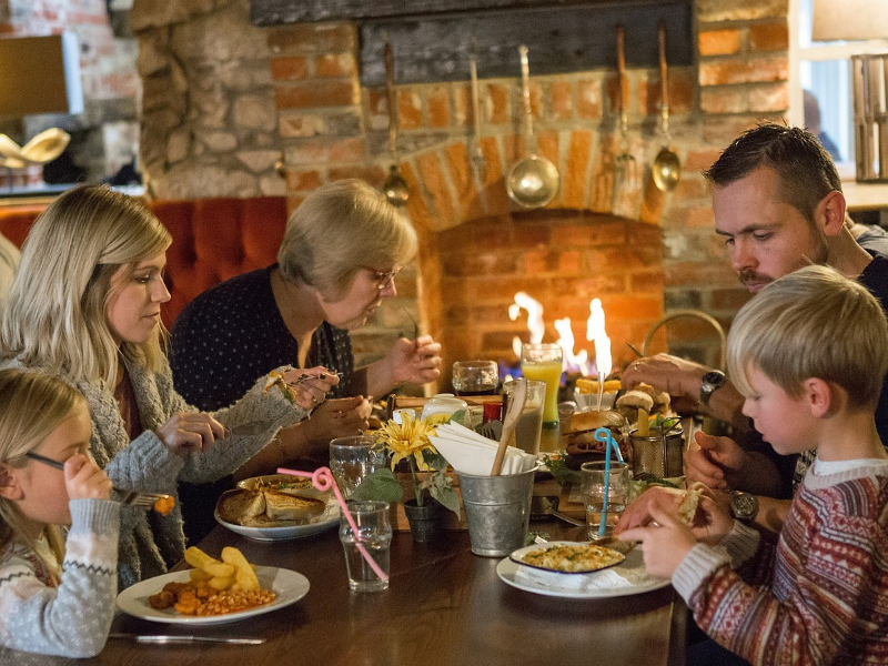 A family of 3 adults and 2 children are dining at a table inside a pub next to a fireplace