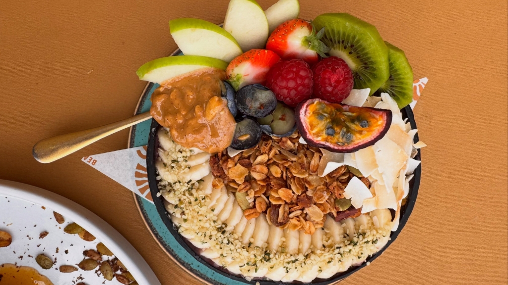 Colourful smoothie bowl topped with fresh fruit, granola, coconut flakes and nut butter, viewed from above on a café table.