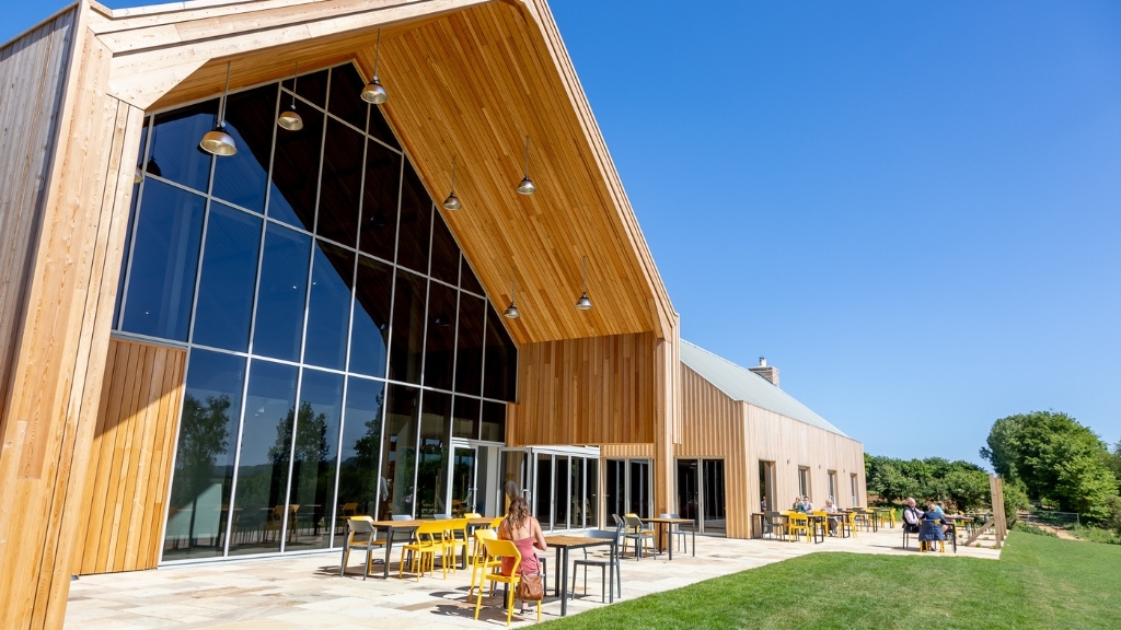 Modern timber-clad farm café with floor-to-ceiling windows and outdoor seating, set beside green fields under a clear blue sky.