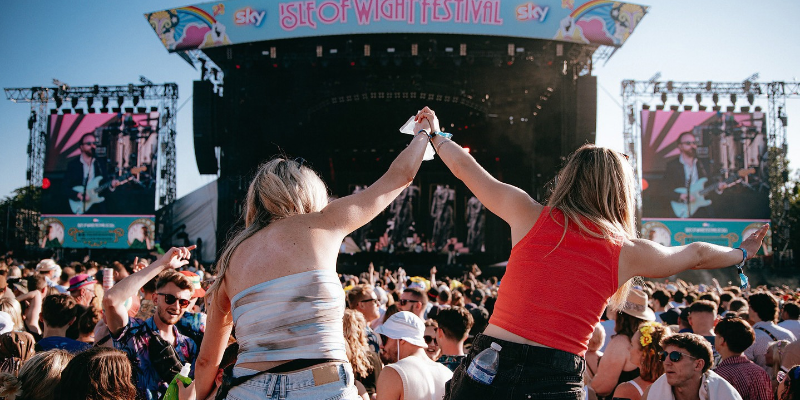 Two women in a crowd at a festival, are on the shoulders of others, and holding hands. In front is the main stage of the Isle of Wight Festival