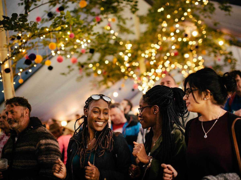 A group of people standing in a festival tent lit with fairy lights, they are laughing and watching the stage