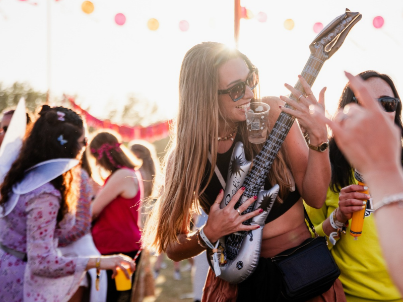 A woman pretends to play an inflatable guitar with a friend next to her. It's sunny and outdoors at a festival