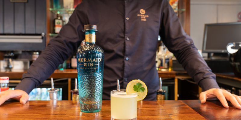 A man in a Mermaid Gin branded black shirt stands behind a bar with a cocktail and bottle of Mermaid gin on it