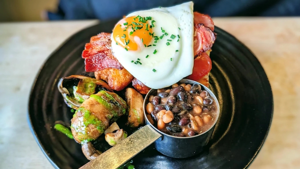 Full cooked breakfast served on a black plate, featuring bacon, a fried egg, roasted vegetables and baked beans in a small metal pot.