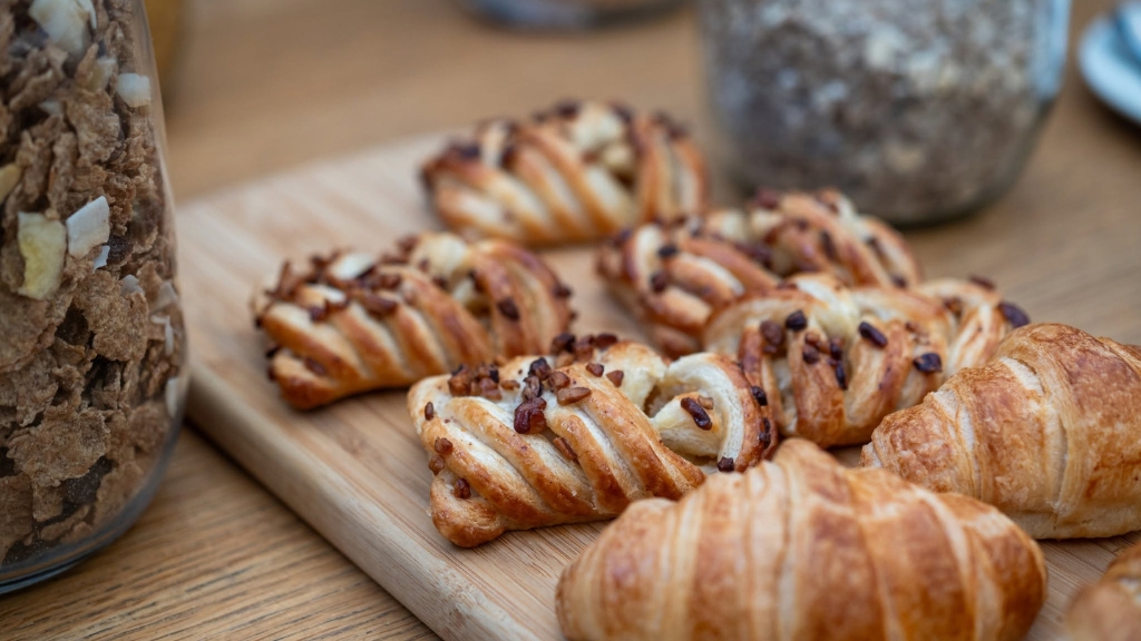 Freshly baked croissants and pastries arranged on a wooden serving board, with jars of breakfast cereals visible in the background.