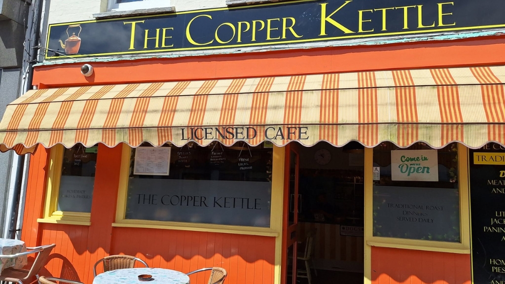 Exterior of The Copper Kettle café, featuring a bright orange frontage, striped awning and outdoor seating on a sunny day.