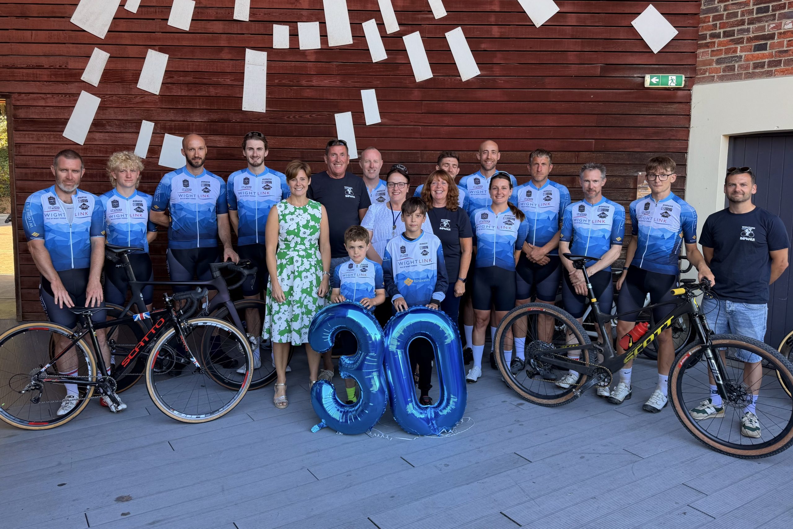 Group photograph of cyclists in blue Wightlink-branded jerseys standing with their bicycles alongside supporters and children, gathered behind large blue "30" balloons in an indoor venue.