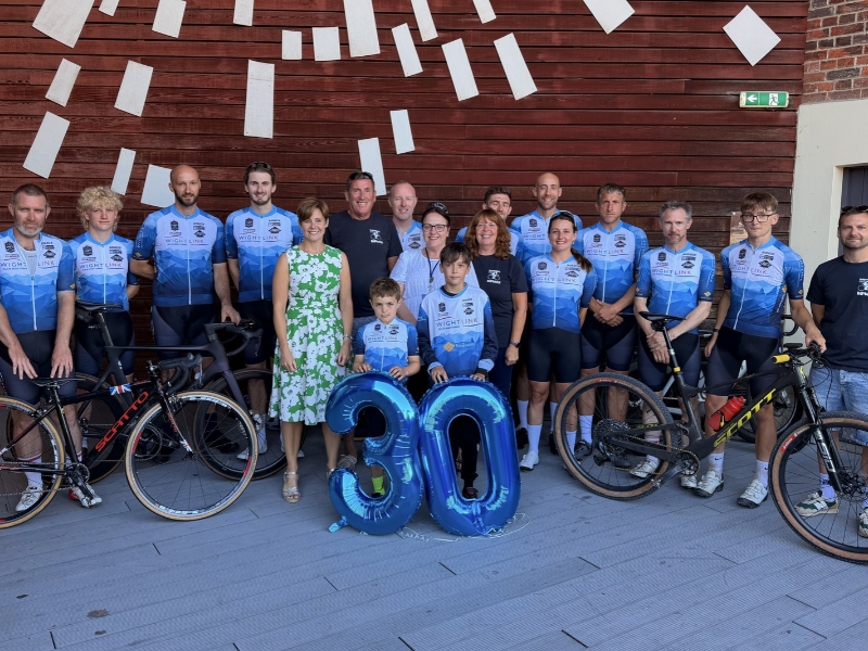 A group of cyclists from the Wightlink Cycle Race Team are assembled at an arts centre with their bikes, with 2 representatives from their sponsor, Wightlink. They have a 3 and a 0 balloon to represent 30 years.
