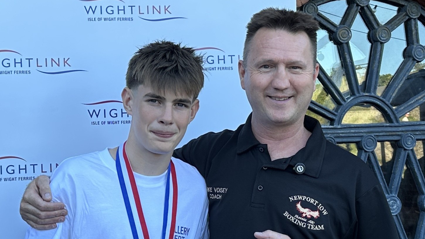Young boxer wearing competition medals stands alongside a coach in front of a Wightlink backdrop following a boxing event.
