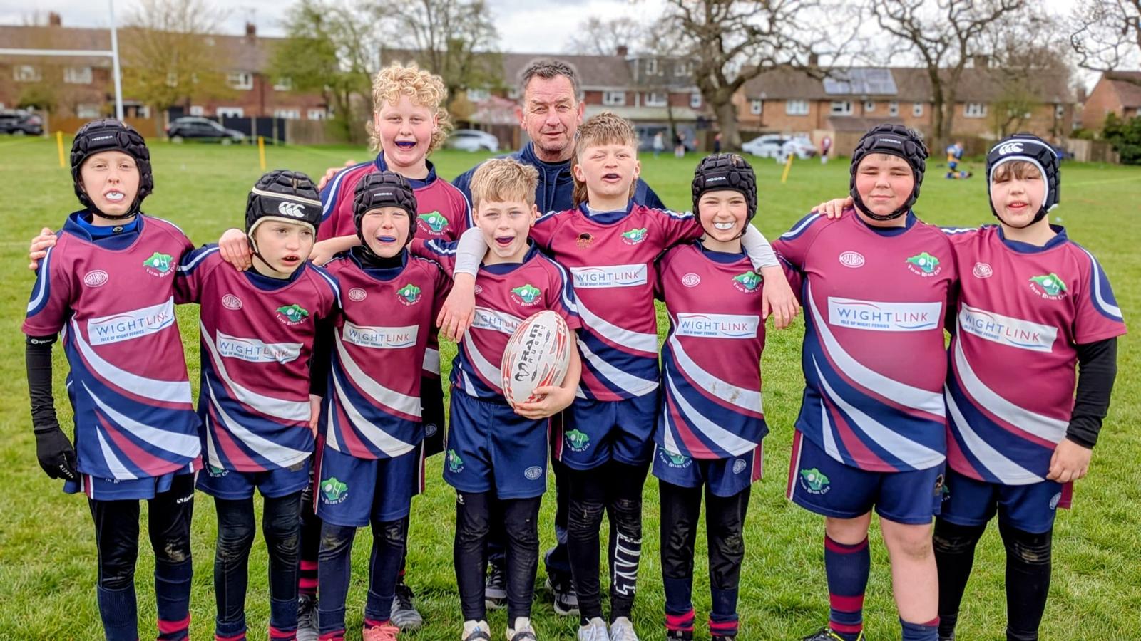 Young players from Vectis Rugby Club pose with their coach on a rugby pitch wearing team kits sponsored by Wightlink. One player holds a rugby ball while teammates stand together ahead of a match or training session.