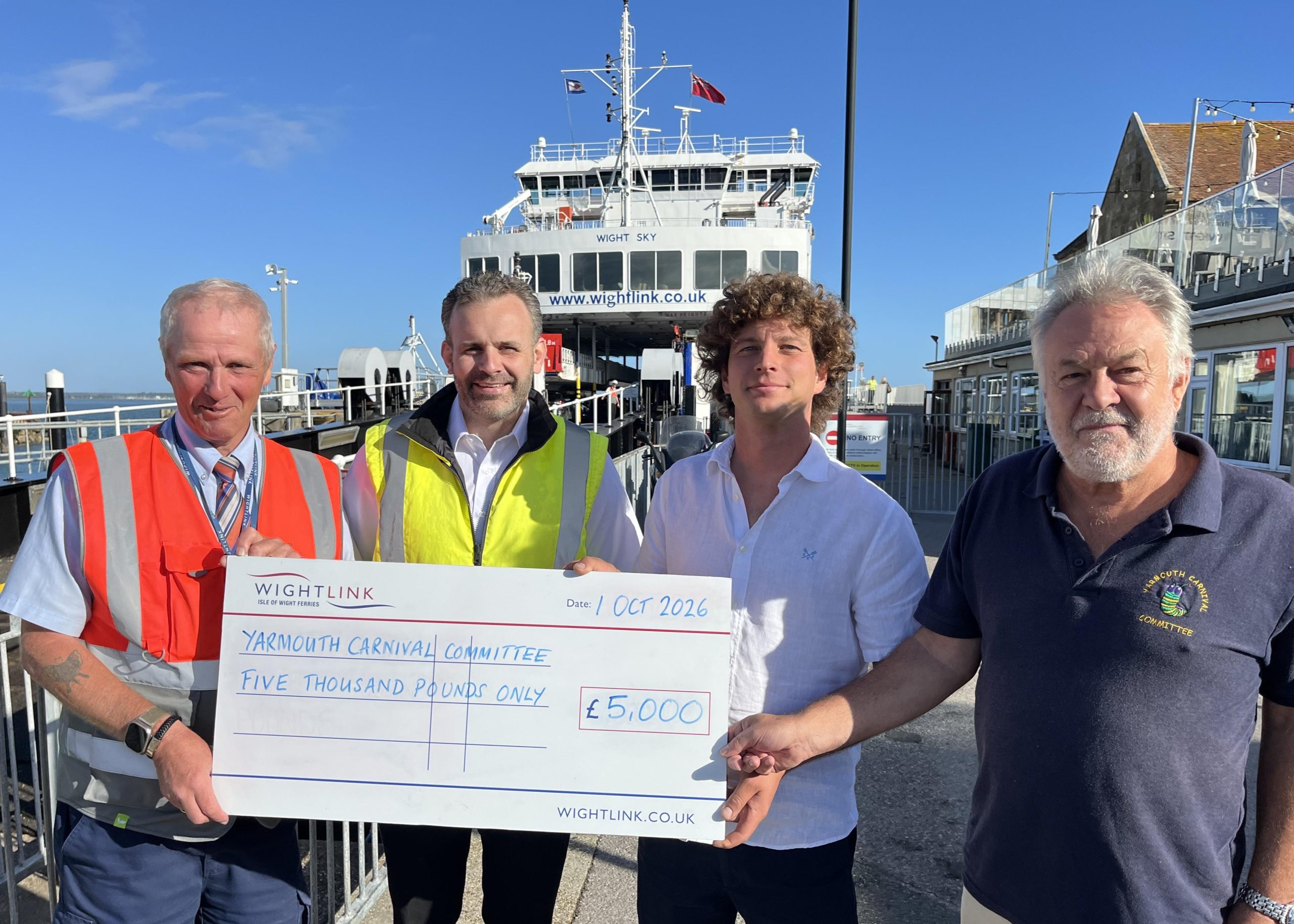 Wightlink representatives and members of the Yarmouth Carnival Committee stand in front of a Wightlink ferry in Yarmouth, holding a ceremonial cheque for £5,000 to support the 2026 Yarmouth Fireworks and Torchlight Procession.