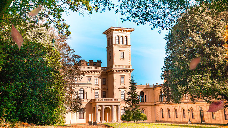 The exterior of a sandstone coloured mansion, Osborne House on the Isle of Wight, framed by foliage